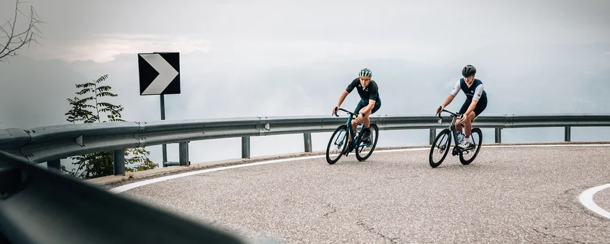 Two cyclists riding on a winding mountain road under cloudy sky