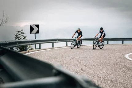 Zwei Radfahrer fahren auf einer kurvigen Bergstraße bei bewölktem Himmel