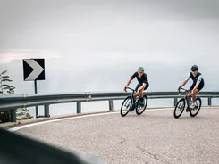 Two cyclists riding on a winding mountain road under cloudy sky
