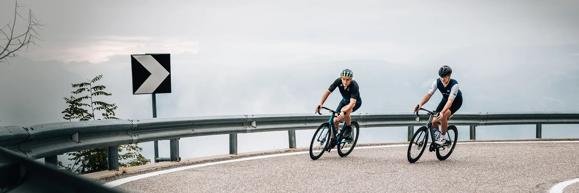 Zwei Radfahrer fahren auf einer kurvigen Bergstraße bei bewölktem Himmel