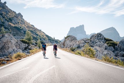 Zwei Radfahrer fahren auf Bergstraße mit Felsen und Gipfeln im Hintergrund