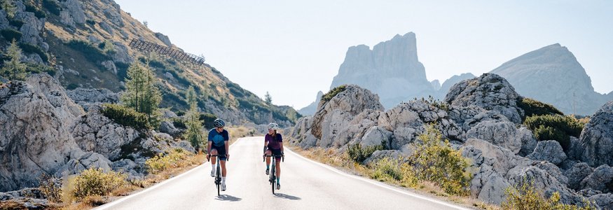 Zwei Radfahrer fahren auf Bergstraße mit Felsen und Gipfeln im Hintergrund