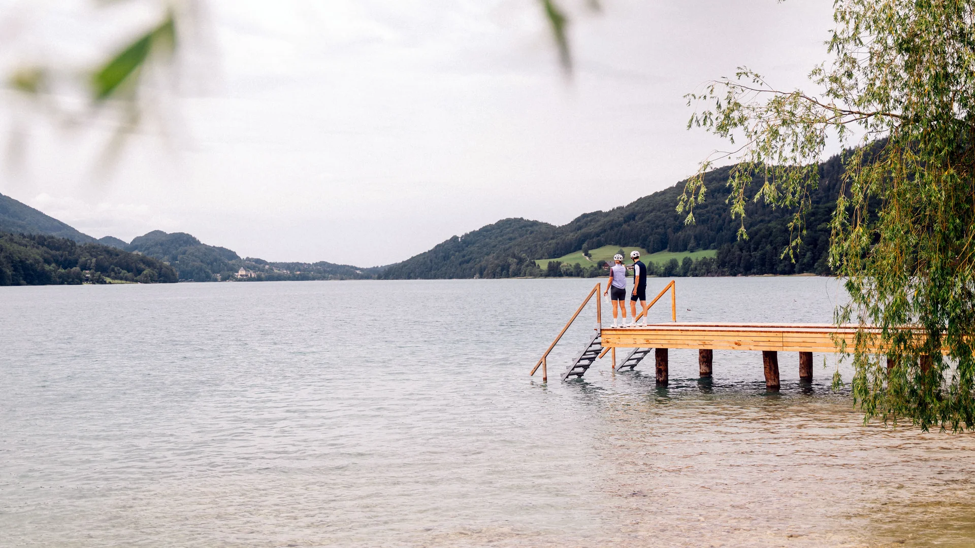 Roadbike Holidays © Stefan Hähnel Zwei Personen stehen auf einem Holzsteg an einem See, umgeben von bewaldeten Hügeln