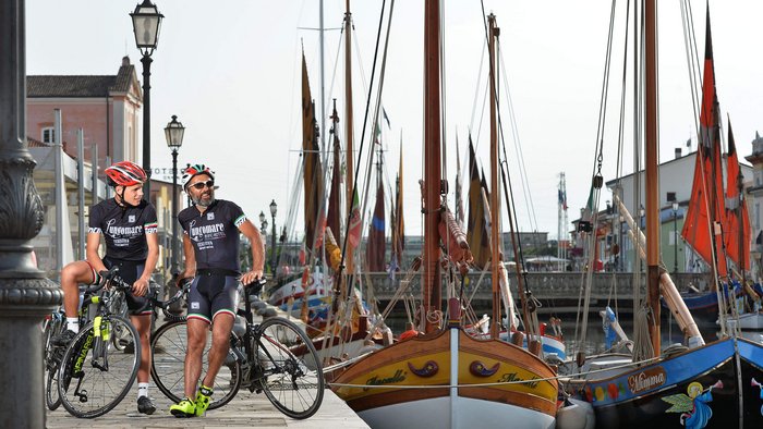 Zwei Radfahrer in Sportkleidung stehen neben Fahrrädern am Hafen mit bunten Booten