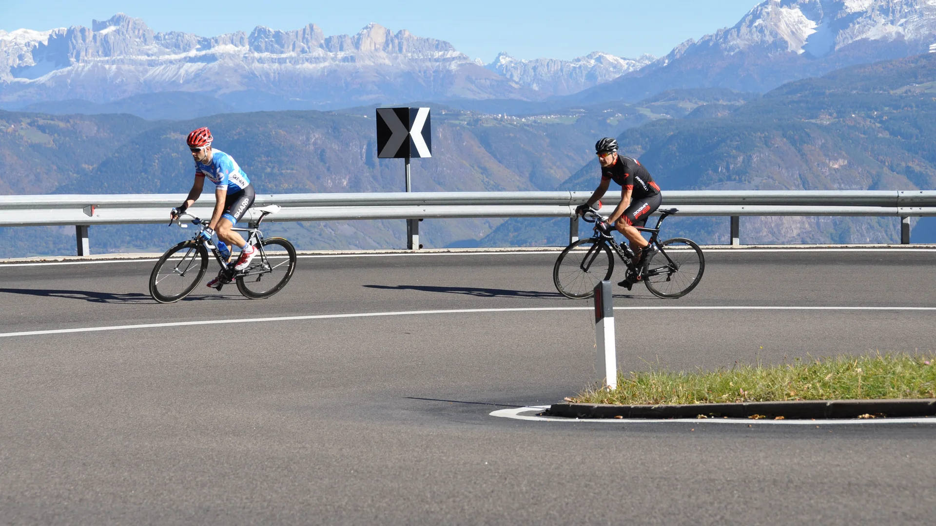 Two cyclists on a winding mountain road with snowy peaks in the background