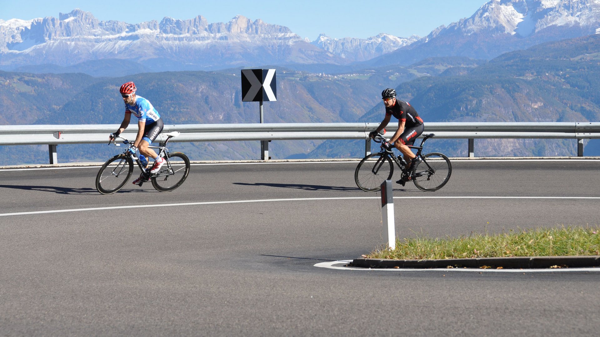 Zwei Radfahrer auf bergiger kurvenreicher Straße mit schneebedeckten Bergen im Hintergrund