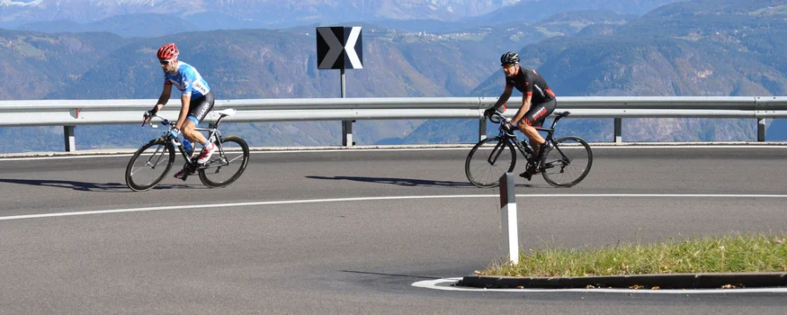 Ein Rennrad-Wochenende in Südtirols Süden © Hotel Teutschhaus Kurtinig Zwei Radfahrer auf bergiger kurvenreicher Straße mit schneebedeckten Bergen im Hintergrund