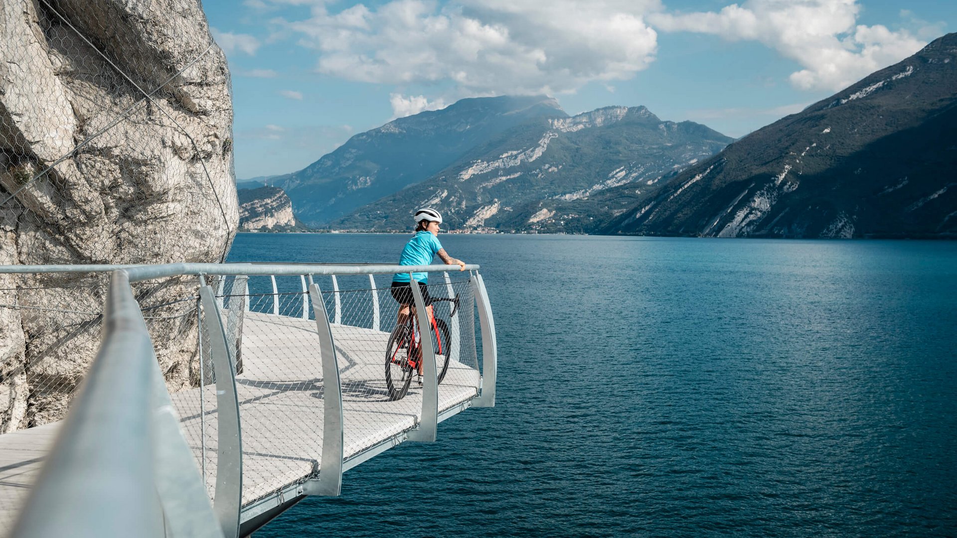 Radfahrer auf Steg mit Blick auf See und Berge bei sonnigem Wetter