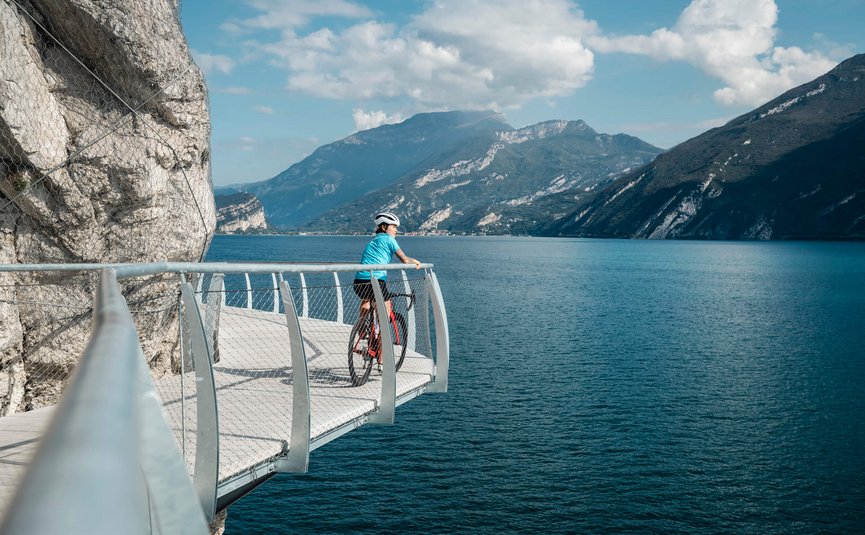 Ciclista su passerella con vista su lago e montagne in una giornata di sole