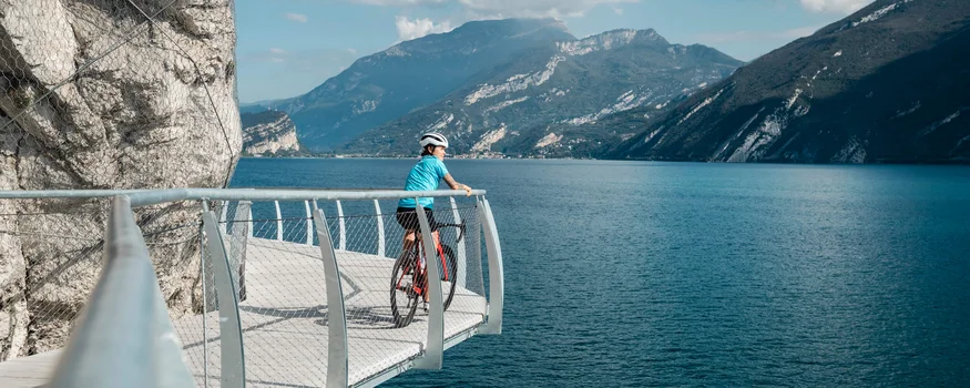 Ciclista su passerella con vista su lago e montagne in una giornata di sole