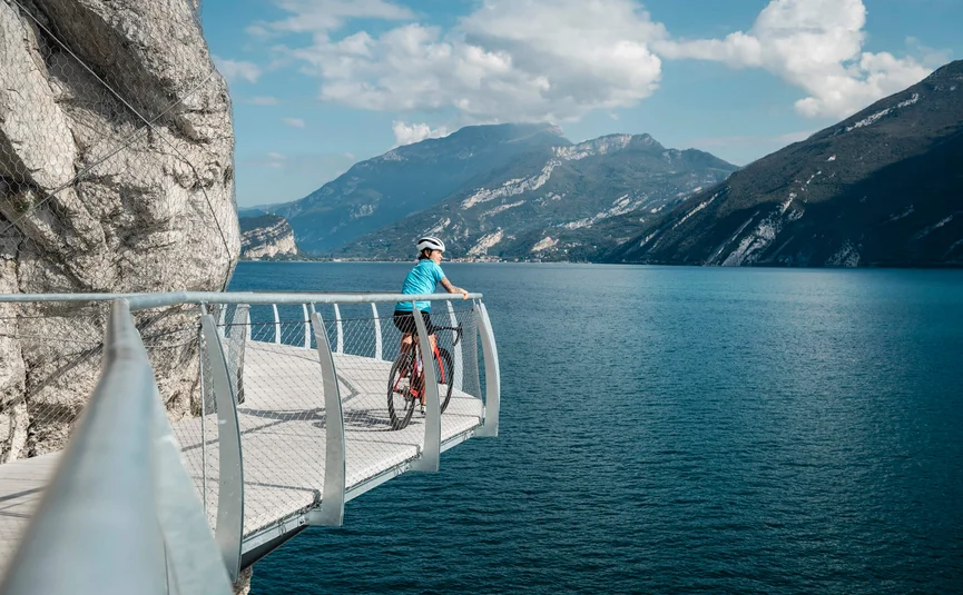 Ciclista su passerella con vista su lago e montagne in una giornata di sole