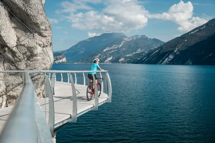 Radfahrer auf Steg mit Blick auf See und Berge bei sonnigem Wetter