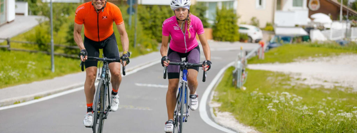 Twee wielrenners in sportkleding rijden op een landweg met huizen op de achtergrond