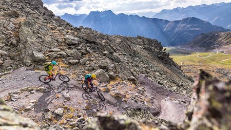 Zwei Mountainbiker fahren auf einem bergigen Pfad mit Alpen im Hintergrund