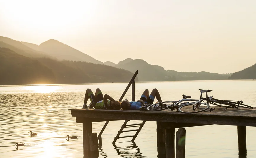 Zwei Radfahrer liegen auf Steg am See im Sonnenuntergang mit Fahrrädern