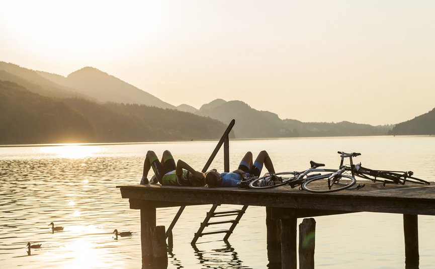Due ciclisti sdraiati su un pontile vicino al lago al tramonto con biciclette