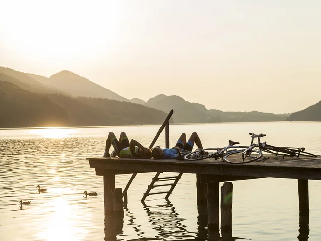 Ein Rennrad-Wochenende im SalzburgerLand – Salzkammergut © SalzburgerLand Fuschlsee - Markus Greber Zwei Radfahrer liegen auf Steg am See im Sonnenuntergang mit Fahrrädern