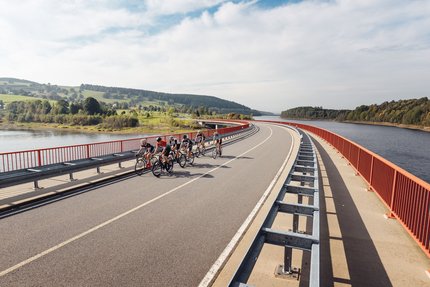 Gruppe von Radfahrern fährt über Brücke neben Fluss und grüner Landschaft