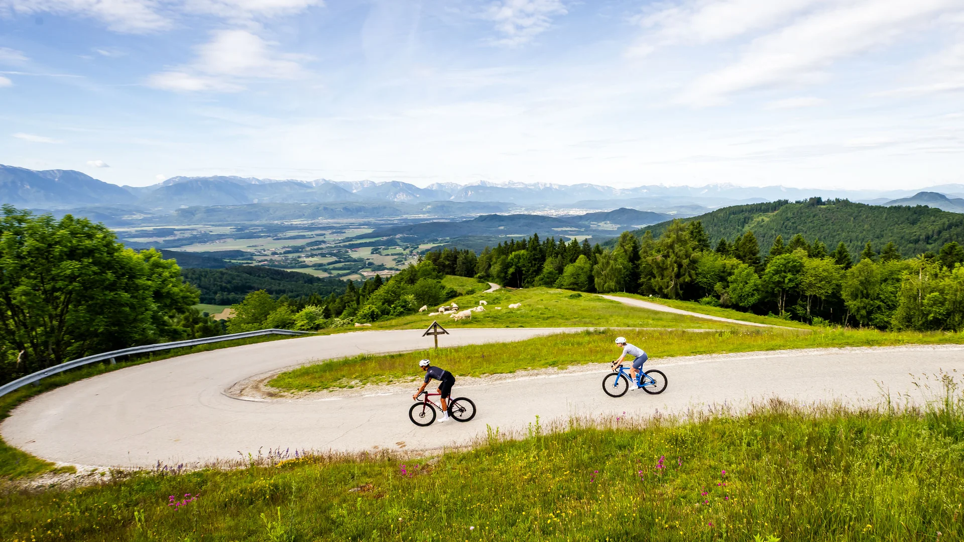 Two cyclists on winding mountain road with green hills and distant mountains