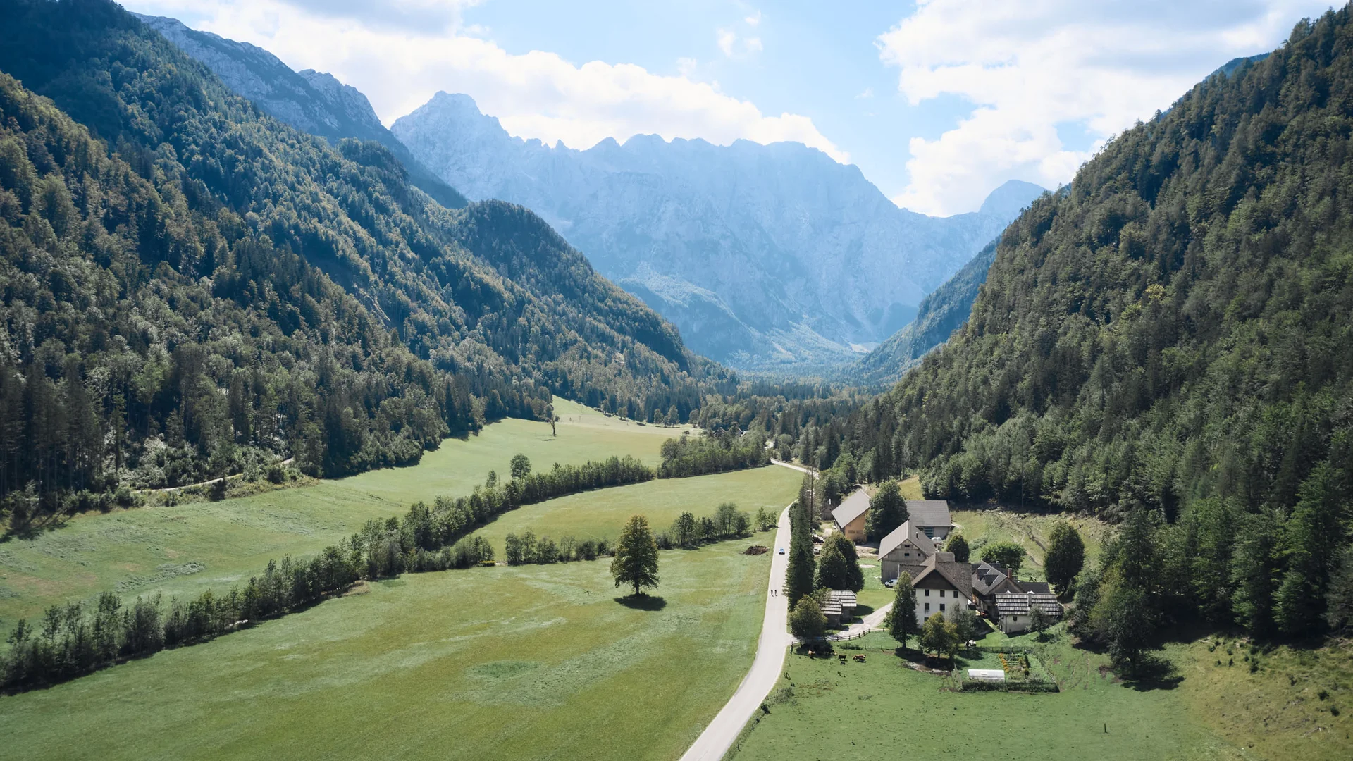 Grünes Tal mit Häusern, bewaldeten Bergen und wolkigem Himmel