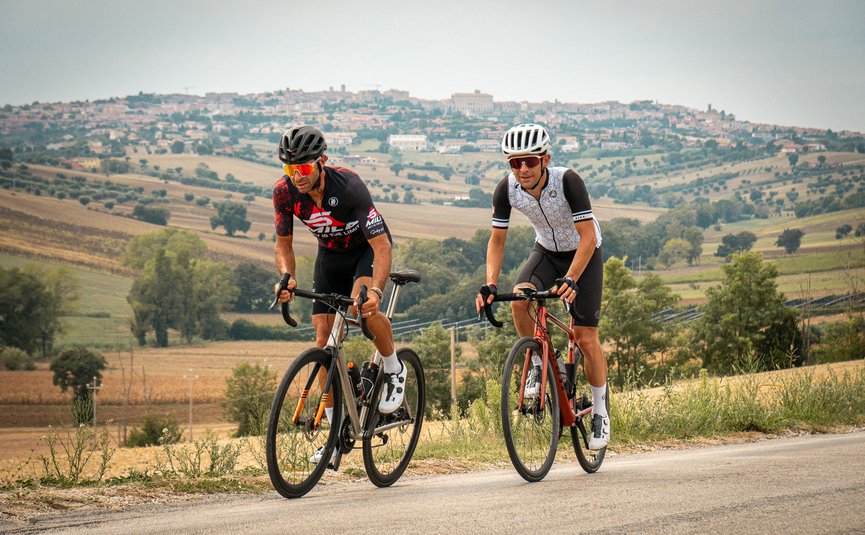 Due ciclisti pedalano su strada di campagna con paesaggio collinare e villaggio sullo sfondo