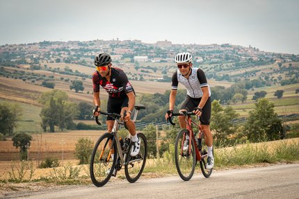 Due ciclisti pedalano su strada di campagna con paesaggio collinare e villaggio sullo sfondo