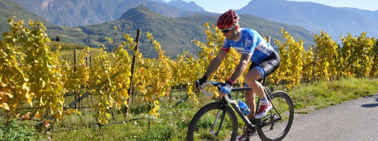 Cyclist riding on country road beside yellow grapevines and mountains under clear sky
