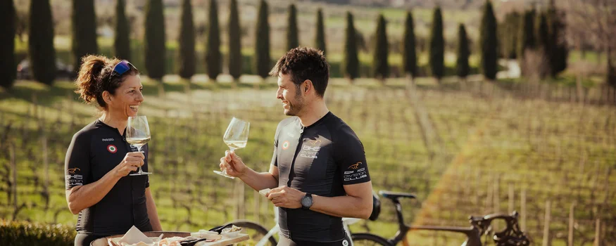 Zwei Radfahrer genießen Wein und Snacks draußen an einem Fass im Weinberg