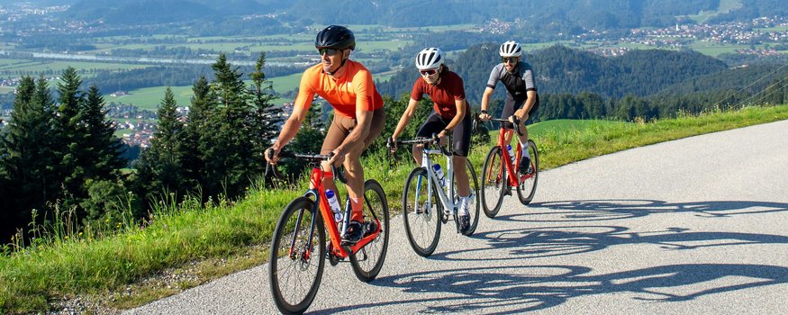 Drei Radfahrer fahren auf einer Bergstraße bei klarem Himmel mit Bergpanorama