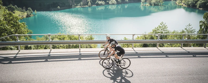 Due ciclisti pedalano su strada vicino a un lago circondato da montagne