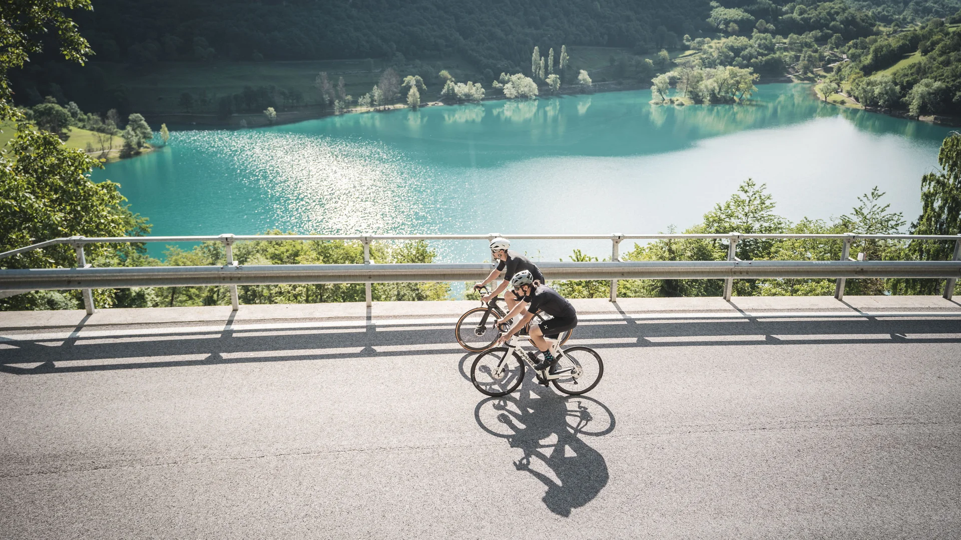 Two cyclists riding on road beside a lake with mountains