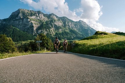 Due ciclisti su strada di montagna con scogliere rocciose e colline verdi sullo sfondo