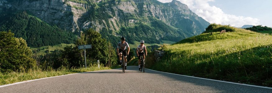 Zwei Radfahrer auf Bergstraße vor beeindruckendem Felsmassiv und grüner Landschaft