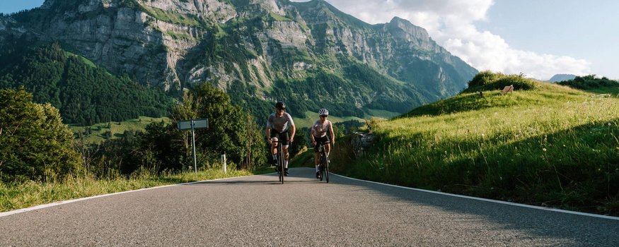 Zwei Radfahrer auf Bergstraße vor beeindruckendem Felsmassiv und grüner Landschaft