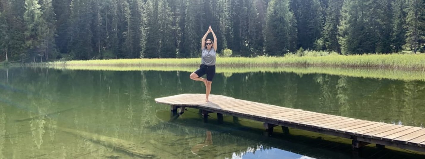 Person macht Yoga auf einem Steg über klarem See mit Wald im Hintergrund