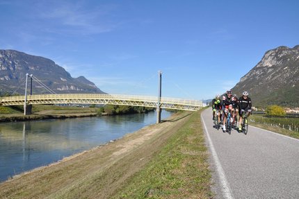 Vijf wielrenners op een weg langs een rivier met brug en bergen