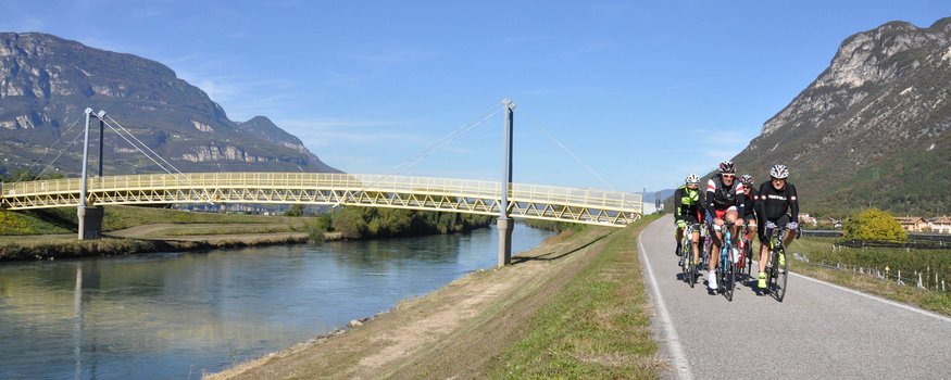 Fünf Radfahrer fahren auf einem Straßenweg entlang eines Flusses mit Brücke und Bergen
