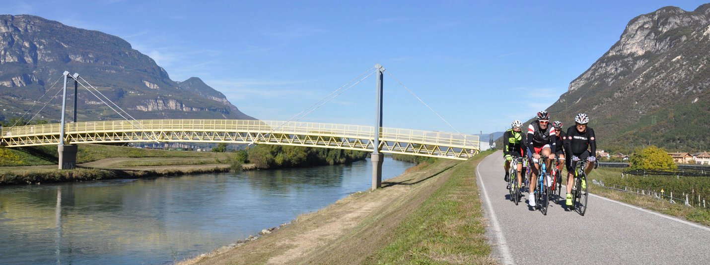 Five cyclists riding on a road next to a river with bridge and mountains