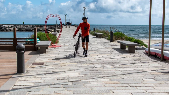 Cyclist walking with bike on a pier by the sea next to a heart decoration