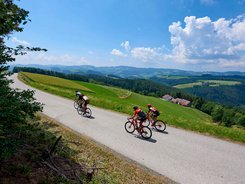 Three cyclists riding on a country road through green hills under a blue sky