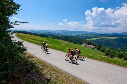 Tre ciclisti su una strada di campagna tra colline verdi sotto un cielo azzurro