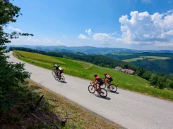 Three cyclists riding on a country road through green hills under a blue sky