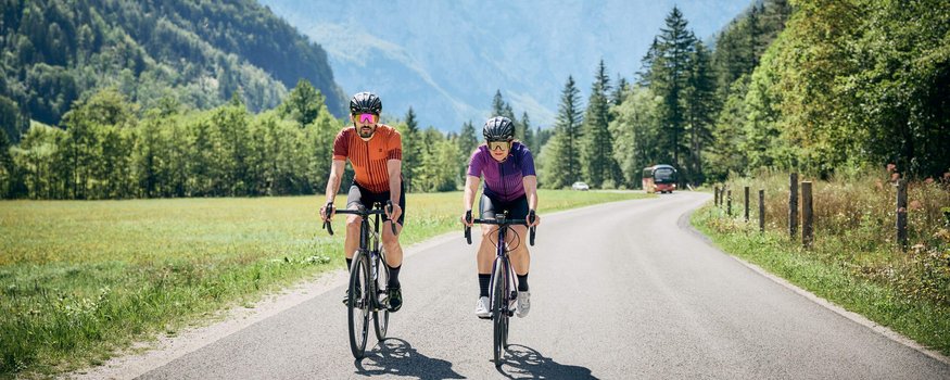 Roadbike + Wellness = perfect Match! © Tobias Köhler Zwei Radfahrer fahren auf einer Landstraße vor Bergen und Wald