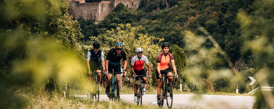 Goldener Herbst auf dem Rennrad in Tramin © Jens Scheibe Vier Radfahrer fahren auf einer kurvigen Straße mit Burg und Wald im Hintergrund