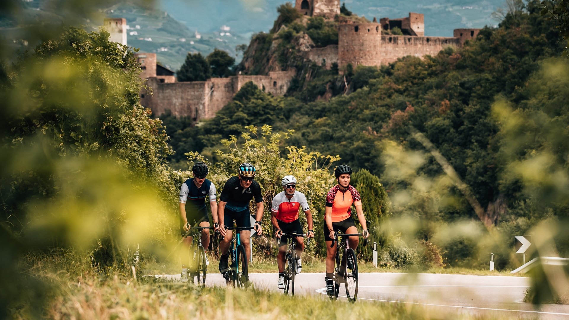 Vier Radfahrer fahren auf einer kurvigen Straße mit Burg und Wald im Hintergrund
