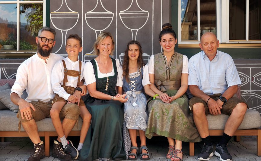 Familie in traditioneller bayerischer Tracht sitzt auf Bank vor Wand mit Glasmuster