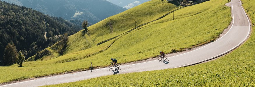 Two cyclists ride a winding mountain road surrounded by green hills