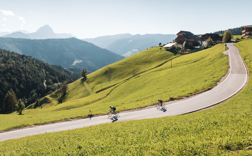 Two cyclists ride a winding mountain road surrounded by green hills