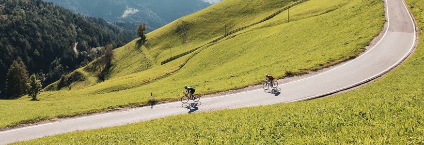 Zwei Radfahrer fahren eine kurvige Bergstraße umgeben von grünen Hügeln
