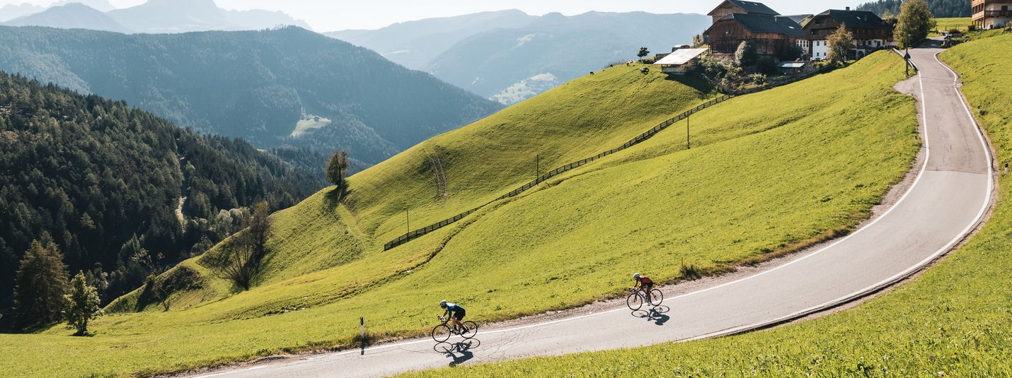 Due ciclisti su una strada di montagna tortuosa circondata da colline verdi